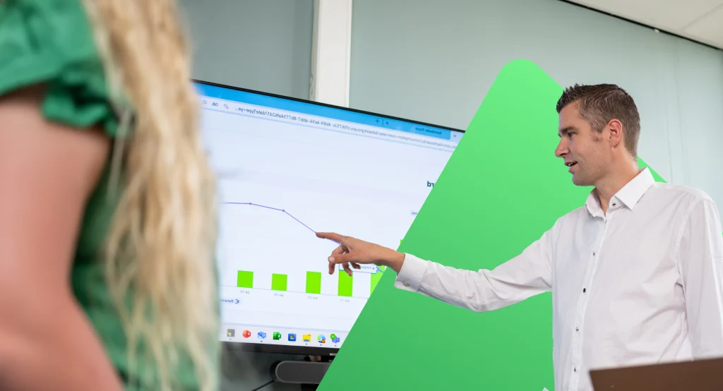 Een man in een witte blouse wijst naar een grafiek op een scherm tijdens een presentatie.