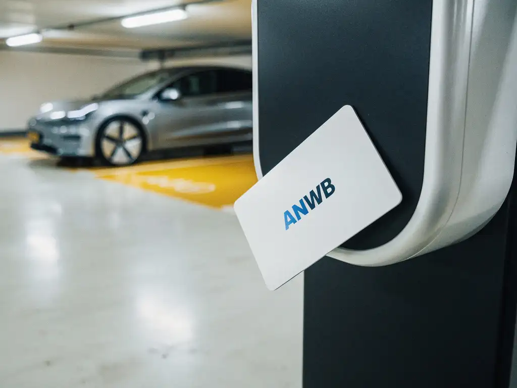 ANWB laadpas leunend tegen een modern EV-laadstation in een parkeergarage, met een elektrische auto op de achtergrond.