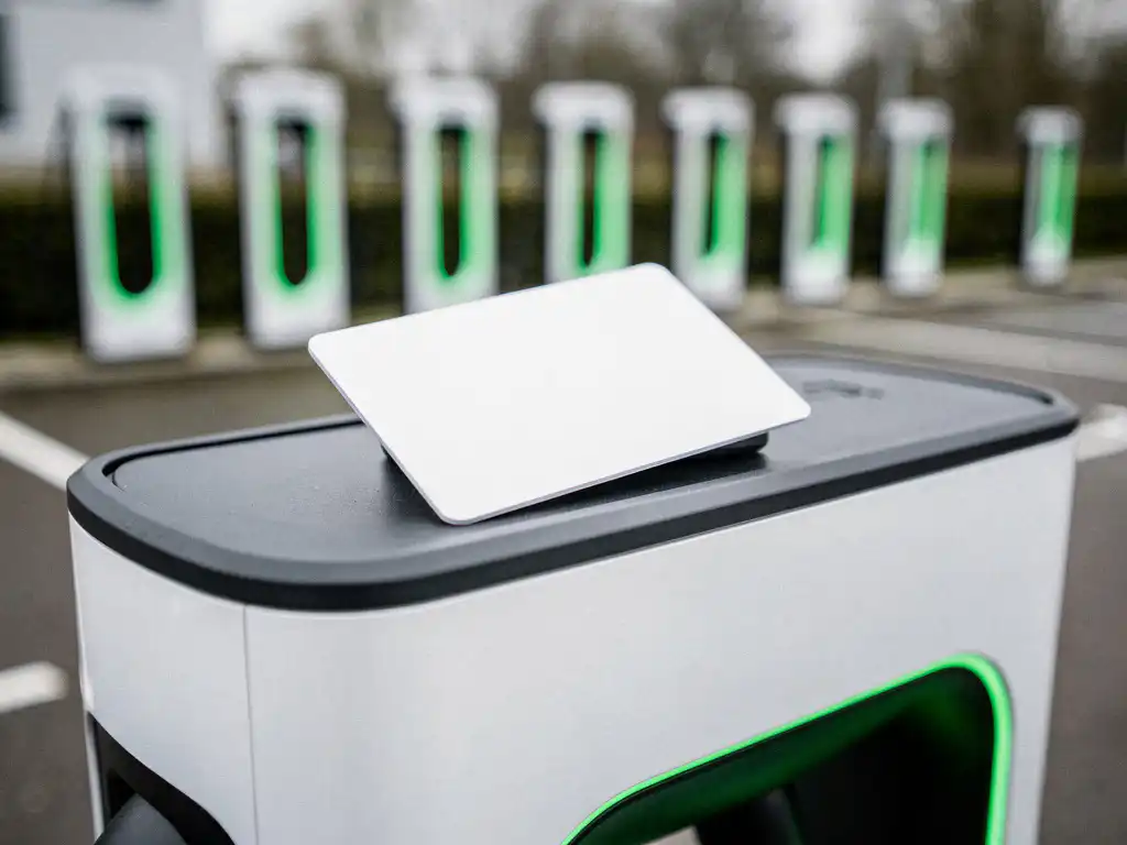 Witte EV-laadpas op een modern laadstation met rij laadpalen op een zakelijk parkeerterrein op de achtergrond.