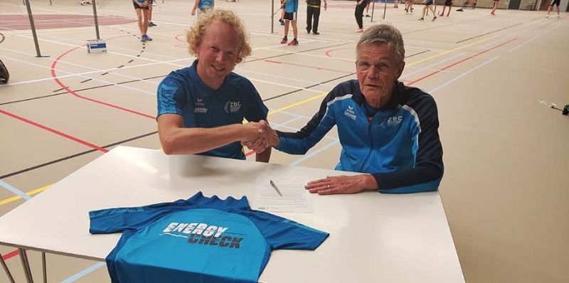 Twee mannen schudden elkaar de hand bij een sponsorcontract met een Energy Check sportshirt op tafel.